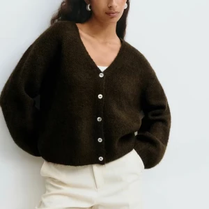 Mörkbrun kofta med v-ringning - Mysig mörkbrun kofta med v-ringning och pärlemorknappar framtill. Koftan har en loose passform och är stickad i ett mjukt material som passar perfekt för kyligare dagar. Långa ärmar och enkel, stilren design.