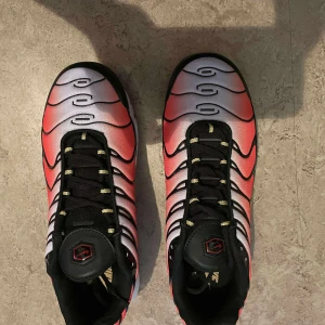 Nike Air Max Plus Tuned röd/svart - Nike Air Max Plus Tuned sneakers med en grym färgskiftning från rött till vitt och svarta detaljer. Snygga vågformade linjer, svart snörning och synlig Air Max-dämpning i sulan. Guldiga detaljer och TN-logga på tungan och sulan. Perfekta för dig som vill sticka ut.