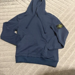 Blå hoodie från Stone Island - Snygg marinblå hoodie från Stone Island med klassisk patch på vänster ärm. Tröjan har huva med dragsko, känguruficka och ribbade muddar. Tillverkad i mjuk bomull, perfekt för en chill och stilren look.