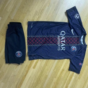 PSG Dembele matchtröja Nike set - Säljer ett PSG matchställ med O. Dembélé #10. Tröjan är mörkblå med röda och grå detaljer, PSG-logga, Qatar Airways-tryck och Champions League-märke. Korta ärmar och klassisk passform. Shorts med PSG-emblem ingår. Perfekt för fotbollsfans!