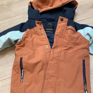 Didriksons vinterjacka orange och blå - Snygg regnjacka från Didriksons i orange, marinblått och ljusblått. Jackan har huva, dragkedja och praktiska fickor med dragkedjor. 100% vattentät och perfekt för blöta dagar. Sportig look med cool färgblock-design och logga på bröstet.