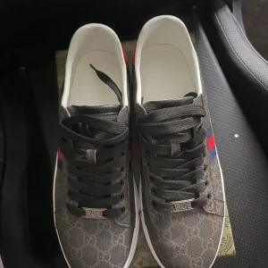Gucci Ace sneakers med monogram - Snygga Gucci Ace sneakers i mörkgrått med klassiskt GG-monogram och röd/blå sidodetalj. Baksidan har röd häl med Gucci-logga. Skorna har svarta snören och platt vit sula. Perfekt för dig som gillar exklusiv streetstyle och vill sticka ut.