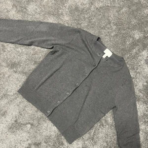 Grå kofta från H&M med knappar - En stilren grå kofta från H&M i storlek S. Koftan är stickad, har rund halsringning och knäpps med matchande knappar framtill. Perfekt att slänga över en t-shirt eller linne för en enkel och clean look.