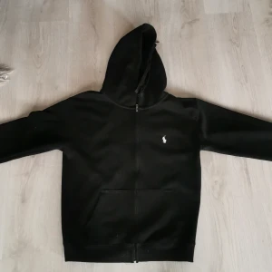 Svart hoodie från Polo Ralph Lauren - Svart hoodie från Polo Ralph Lauren med klassisk vit broderad logga på bröstet. Tröjan har dragkedja framtill, huva med snörning och känguruficka. Tillverkad i mjuk bomullsmix, perfekt för chill dagar eller när du vill ha en clean streetstyle.