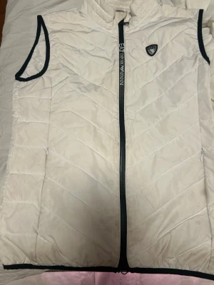Vit dunväst från Emporio Armani EA7 - Säljer en snygg vit dunväst från Emporio Armani EA7. Västen har svarta detaljer vid ärmöppningarna och dragkedjan, samt ett EA7-märke på bröstet. Quiltad design och två fickor med dragkedja. Perfekt för lager-på-lager och streetwear. Den har några fläckar men det går nog bort i tvätten 