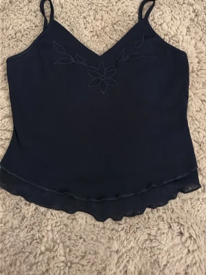 Svart linnetopp med broderad blomma - Säljer en svart linnetopp med tunna axelband och v-ringning. Toppen har en broderad blomma framtill och vågig kant nertill för en extra detalj. Materialet känns lätt och luftigt, perfekt för varma dagar.