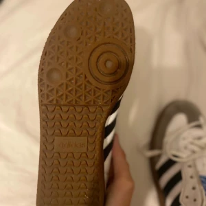 Adidas Samba sneakers og - Adidas Samba sneakers med klassisk design, vita med grå mockadetaljer och svarta ränder på sidorna. Skorna har snörning, platt gummisula och rund tå. Märkets blå logga syns på tungan. Perfekt för dig som gillar retrostil och sportig vibe. Har bara använt som inomhus skor på idrott. Aldrig utomhus.
