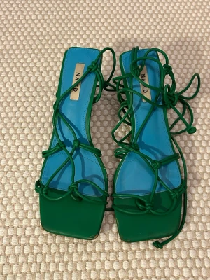Gröna sandaletter med snörning från NA-KD - Säljer ett par trendiga sandaletter från NA-KD i klargrönt med blå innersula. Skorna har fyrkantig tå, tunna snören som går att knyta runt benet och en låg klack. Tillverkade i syntetmaterial och perfekta för sommarens outfits. Se matchande väska i annat innehåll
