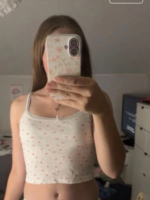 Vit croppad linne med rosettprint Gina Tricot - Supersöt croppad linne  från Gina Tricot i vit bomull med tunna axelband och små rosa rosetter som mönster. Linnet  har spetskant upptill och en liten rosett i mitten. Perfekt för en myskväll!