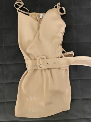 Beige satin topp med bälte - Snygg beige satin topp från In The Style med justerbara tunna axelband och matchande bälte i midjan som också går att justera. Toppen har en omlottdesign och är tillverkad i ett mjukt, lätt tyg som känns skönt mot huden. Perfekt för dig som gillar stilrena och trendiga plagg. Helt ny. Angående frågor är det bara att höra av sig, samma gäller även diskussioner kring priset. 