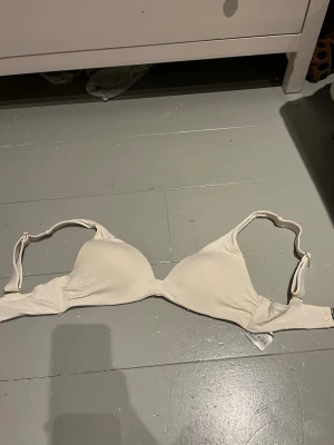 Vit ribbad bikiniöverdel från H&M - Säljer en vit bikiniöverdel från H&M i storlek 38. Överdelen har ribbad struktur, justerbara axelband och guldfärgade detaljer vid banden. Enkel och stilren design som passar perfekt till sommaren.