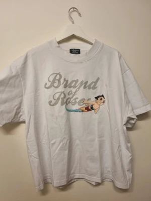 Vit Brand of Roses t-shirt med tryck - Säljer en vit t-shirt från Brand of Roses i storlek L. T-shirten har ett stort tryck framtill med texten 'Brand of Roses' och en tecknad figur. Klassisk rund hals och korta ärmar. Tillverkad i mjuk bomull för skön känsla.