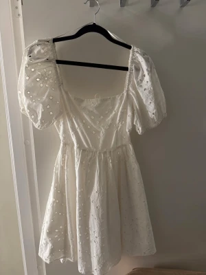 Vit kort klänning med puffärm - Supersöt vit kort klänning med puffärmar och broderade detaljer. Klänningen har fyrkantig ringning och är gjord i bomull. Perfekt till studenten och sommaren ☺️