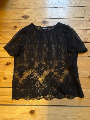 Svart spetsblus från Pieces - Säljer en transparent svart blus från Pieces med vertikala ränder och vacker spetsdetalj nertill. Blusen har rund halsringning och korta ärmar. Perfekt för dig som gillar en edgy och feminin stil. Priset kan diskuteras. Skriv vid frågor😊