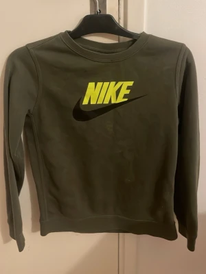 Grön Nike sweatshirt med logga - Säljer en grön Nike sweatshirt i storlek L junior med stor gul och svart Nike-logga på bröstet. Tröjan har rund halsringning och långa ärmar. Perfekt för dig som gillar sportig och enkel stil.