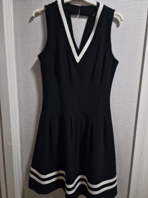 Svart kortklänning med vita detaljer H&M - Supersnygg svart kortklänning från H&M med vita ränder vid fållen och V-ringad hals med vit kant. Klänningen är ärmlös och har en markerad midja samt dragkedja i ryggen. Perfekt för dig som gillar sportig och stilren look.