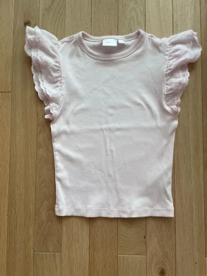 Ljusrosa topp med volangärmar - Supersöt ljusrosa topp från Gina Tricot med ribbad struktur och breda volangärmar i broderad bomull. Ärmarna har fina spetsdetaljer och ger en romantisk vibe. Perfekt till jeans eller kjol för en söt look.