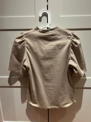 Beige topp med puffärm - Säljer en beige topp med puffärm och hög hals. Toppen har korta ärmar och är gjord i ett mjukt material som känns skönt mot huden. Enkel men ändå trendig design som passar till mycket. Perfekt för dig som gillar stilrena plagg med en twist.
