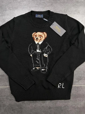 Svart stickad tröja Polo Ralph Lauren - Svart stickad tröja från Polo Ralph Lauren med ikonisk björn i kostym broderad på bröstet. Rund halsringning och ribbade muddar vid ärmslut och nederkant. RL-broderi nere vid fållen. Perfekt för dig som vill ha en klassisk men lekfull look.