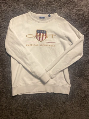 Vit sweatshirt från GANT - Vit sweatshirt från GANT med broderad logga och texten 'American Sportswear' framtill. Tröjan har rund halsringning, långa ärmar och ribbade muddar. Klassisk och stilren design med detaljer i guld och vit 