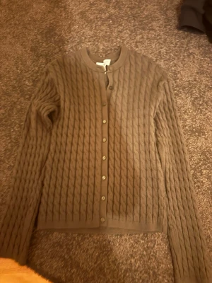 Mörkgrön kabelstickad kofta med knappar - Säljer en mörkgrön kabelstickad kofta med rund hals och knäppning framtill. Koftan har lång ärm och ett klassiskt mönster som ger en mysig och stilren look. Perfekt att slänga på sig när det är kyligt ute.