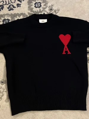 Svart stickad tröja från Ami Paris - Svart stickad tröja från Ami Paris med röd hjärtlogga och bokstaven A på bröstet. Tröjan har ribbade muddar och klassisk passform med rund hals. Perfekt för dig som gillar stilrena plagg med en twist. Skön, snygg, storlek S 160-170