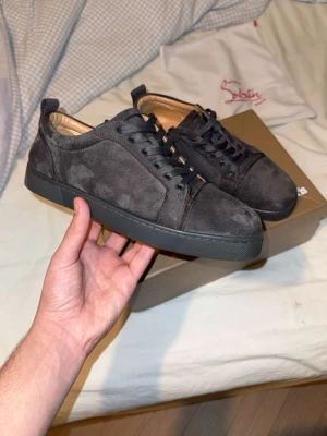 Mörkgrå mocka sneakers från Louboutin - Säljer ett par stilrena mörkgrå sneakers från Christian Louboutin i mjuk mocka. Skorna har rund tå, svarta snören och diskret svart sula. Insidan är fodrad i beige skinn för extra komfort. Perfekta för dig som gillar lyxiga detaljer och klassisk design.