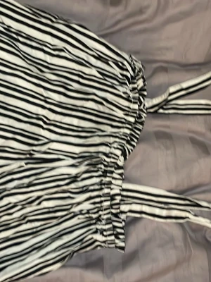 Randig svartvit linnetopp med knytband - Säljer en snygg randig linnetopp i svart och vitt med smala axelband som knyts upptill. Toppen har resår vid bysten för en bekväm passform och är gjord i ett mjukt, lätt material. Perfekt för varma dagar och enkel att styla med jeans eller kjol.