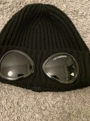 Svart mössa med goggles från C.P. Company - Säljer en svart ribbstickad mössa från C.P. Company. Mössan är äkta och den är i storlek M