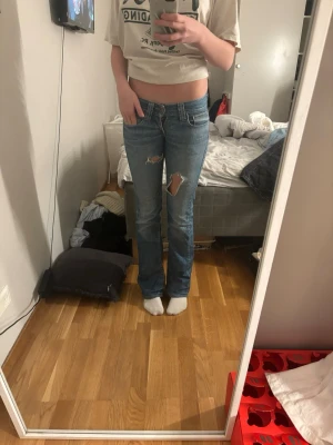 Blå bootcut jeans med slitningar - Säljer ett par blå bootcut jeans med slitningar på låret och vid fickan. Jeansen har låg midja och klassisk femficksdesign. Materialet är denim och passformen är normal med lätt utsvängda ben. Perfekta för en avslappnad och trendig look.