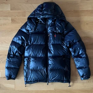 Rhalp Laurent pufferjacka  - Säljer en mörk blå pufferjacka från Ralph Lauren med röd broderad logga på bröstet. Jackan har huva, dragkedja framtill och flera fickor. Materialet är glansigt och känns riktigt varmt, perfekt för kalla dagar. Den har en liten defekt vid handleden och en liten defekt på insidan av jackan med är Lagad så inga större problem med det annars jätte gott skick. Pris kan diskuteras. 