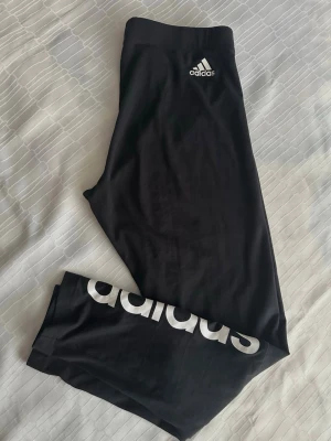Svarta leggings från Adidas S - Säljer ett par svarta leggings från Adidas med hög midja och stor vit Adidas-logga längs ena benet. Tight passform och stretchigt material som passar perfekt till träning eller chill. Klassisk look med diskret logga vid höften och bred resår i midjan.