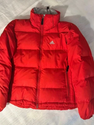Röd dunjacka från Adidas storlek 38, dun 80%, fjäder 20%.  - Säljer en röd pufferjacka från Adidas med hög krage och grått innerfoder. Jackan har dragkedja framtill, två sidofickor med dragkedja och ett stilrent Adidas-tryck på bröstet. Perfekt för kalla dagar och har en riktigt schysst streetstil. 