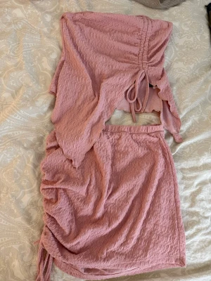 Topp med kjol sett - Säljer en rosa croppad väst i ett strukturerat, lätt skrynkligt tyg. Västen har snörning framtill och en avslappnad passform. Perfekt för dig som vill ha en trendig och bekväm look.