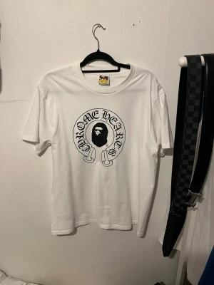 Vit Chrome Hearts x BAPE t-shirt - Säljer en clean vit t-shirt från Chrome Hearts x A Bathing Ape med stor svart logga och ikoniskt ape-motiv på bröstet. På ryggen finns Baby Milo-grafik och Chrome Hearts-tryck. Klassisk passform och crewneck. Perfekt för dig som gillar streetwear och unika collabs.