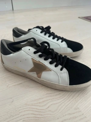 GGDB Superstar sneakers med stjärna - Säljer ett par GGDB Superstar sneakers i vitt skinn med svart tå och svarta skosnören. Sidan har en ikonisk guldbrun stjärna och GGDB/SSTAR-tryck. Skorna har rund tå, platt sula och svart detalj på hälen. Perfekta för dig som gillar streetstyle och vill ha något unikt.