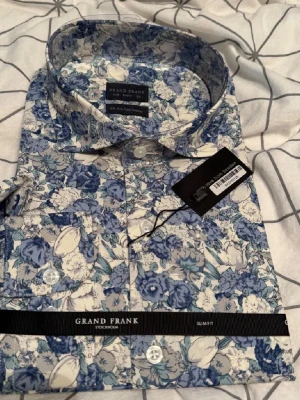 Blommig skjorta från Grand Frank - Säljer en oanvänd skjorta från Grand Frank med blått och vitt blommönster. Skjortan har klassisk krage, vita knappar och är perfekt för dig som vill sticka ut med en snygg och färgstark look. Storlek är L och passform är slim fit.