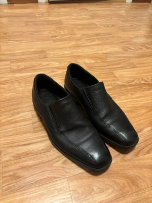 Svarta loafers från Ecco i skinn - Säljer ett par klassiska svarta loafers från Ecco i mjukt skinn. Skorna har en stilren design med rund tå, låg klack och elastiska partier på sidorna för enkel påtagning. Perfekta till både jeans och kostym för en clean look.