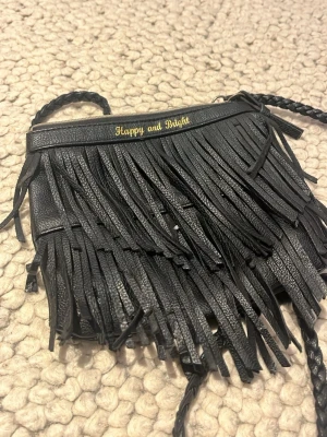 Svart kuvertväska med fransar - Säljer en svart kuvertväska från Happy and Bright med långa fransar framtill och flätad axelrem. Väskan är i skinnimitation och har dragkedja upptill. Insidan är ljud. Perfekt för dig som vill sticka ut med en cool accessoar.