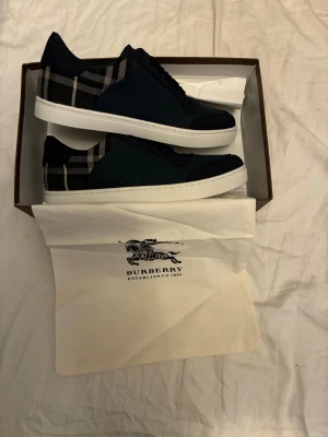 Burberry sneakers i blått - Snygga Burberry sneakers med klassiskt rutigt mönster på hälen och mörkblå ovandel. Skorna har vit platt sula med Burberry-logga, snörning och detaljer i skinn och textil. Perfekt för dig som gillar stilrena och lyxiga sneakers med unik design.