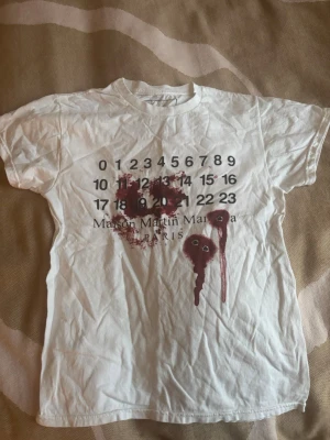 Maison Martin Margiela vit t-shirt S - Lite skrynklig i fotot med et är bara stryka Unik vit t-shirt från Maison Martin Margiela med ikoniskt kalendertryck och röda färgstänk framtill. T-shirten är i 100% bomull och har en klassisk passform med korta ärmar och rund hals. Perfekt för dig som gillar statement-plagg och streetwear-vibe.