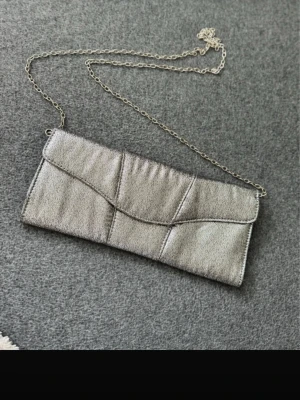 Silvrig kuvertväska med kedja från dipp - Snygg silvrig kuvertväska från dipp med kedjerem och magnetlås. Väskan har ett metallicskimrande yttertyg och flera fack inuti för småsaker. Perfekt när du vill addera lite extra bling till din outfit.