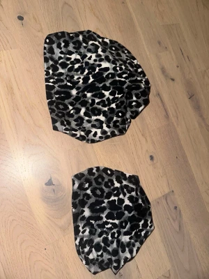 Leopardmönstrad väst i stretch Matchande Huvudbonader i Leopardmönster - Set om 2 - Ett set med två matchande huvudbonader i ett svartvitt leopardmönster. Den ena är i större storlek och den andra i mindre. Säljer en leopardmönstrad väst i svart, grått och vitt. Västen har en cool omlott-look och är tillverkad i ett tunt syntetmaterial. Perfekt för dig som vill sticka ut med ett djurmönster och addera lite edge till din outfit. En vuxen och en för barn cirka 6 mån +