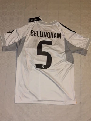 Real Madrid Bellingham matchtröja - Säljer en officiell Real Madrid matchtröja med Bellingham och nummer 5 på ryggen. Tröjan är vit med grå och gula detaljer, svarta Adidas-ränder på axlarna och klubbmärke på bröstet. Tillverkad i lätt och ventilerande polyester, perfekt för fotboll.