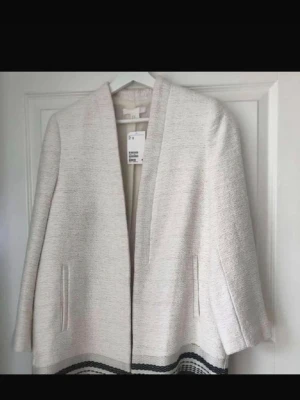 Beige kappa med svarta detaljer - Stilren beige kappa från H&M med raka linjer och öppen front. Jackan har diskreta fickor och svarta ränder längst ner som ger en snygg kontrast. Perfekt för dig som gillar en clean och modern look. Inte använd