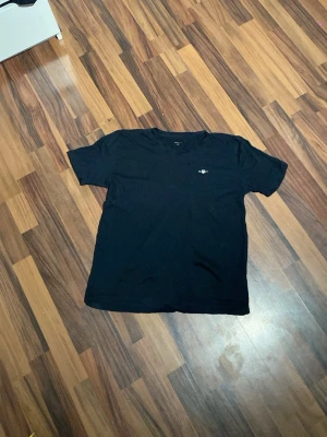 Svart t-shirt från GANT - En svart t-shirt från GANT med klassisk passform och rund hals. På bröstet finns en liten broderad GANT-logga med amerikansk sköld. Tillverkad i mjuk bomull som känns skön mot huden. Perfekt till jeans eller shorts.