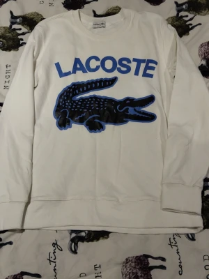 Vit sweatshirt från Lacoste med blå logga - Säljer en clean vit sweatshirt från Lacoste med stor blå logga och ikonisk krokodil på bröstet. Tröjan har rund halsringning och långa ärmar. Perfekt för dig som gillar streetwear och vill ha något stilrent men ändå med statement.