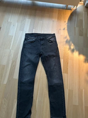 Svarta jeans från Nudie Jeans 32/32 - Säljer ett par svarta jeans från Nudie Jeans i storlek 32/32. Klassisk femficksmodell med raka ben och normal passform. Jeansen är tillverkade i ett slitstarkt bomullstyg och har en stilren look som funkar till det mesta. Passformen är Lean Dean vilket är som slim fit. Skicket är väldigt bra kan inte se att de är använda, nypris 1600kr