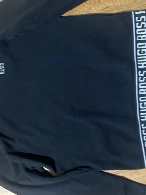 Svart sweatshirt från Hugo Boss - Svart sweatshirt från Hugo Boss med vit logga broderad på bröstet och bred resår med Hugo Boss-tryck längst ner. Klassisk rund halsringning och långa ärmar. Perfekt för en clean och stilren look.
