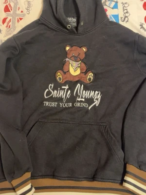 Svart hoodie med björn från Sainte Youngz - Svart hoodie från Sainte Youngz med en stor söt björn på bröstet och texten 'Trust Your Grind'. Hoodien har känguruficka och ribbade muddar med bruna och beige ränder. Perfekt för dig som gillar streetwear och vill ha en unik look.